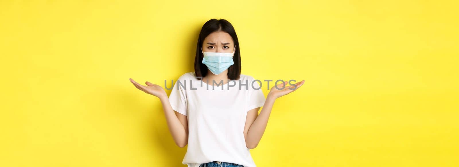Covid-19, quarantine and social distancing concept. Confused asian woman in medical mask shrugging shoulders, spread hands sideways clueless, know nothing, yellow background by Benzoix