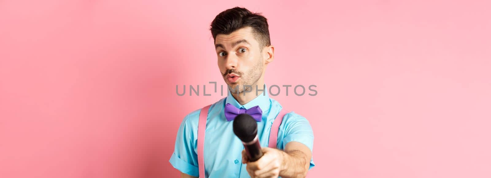 Handsome male show host stretch out hand with microphone, asking for interview or comments, standing on pink background by Benzoix