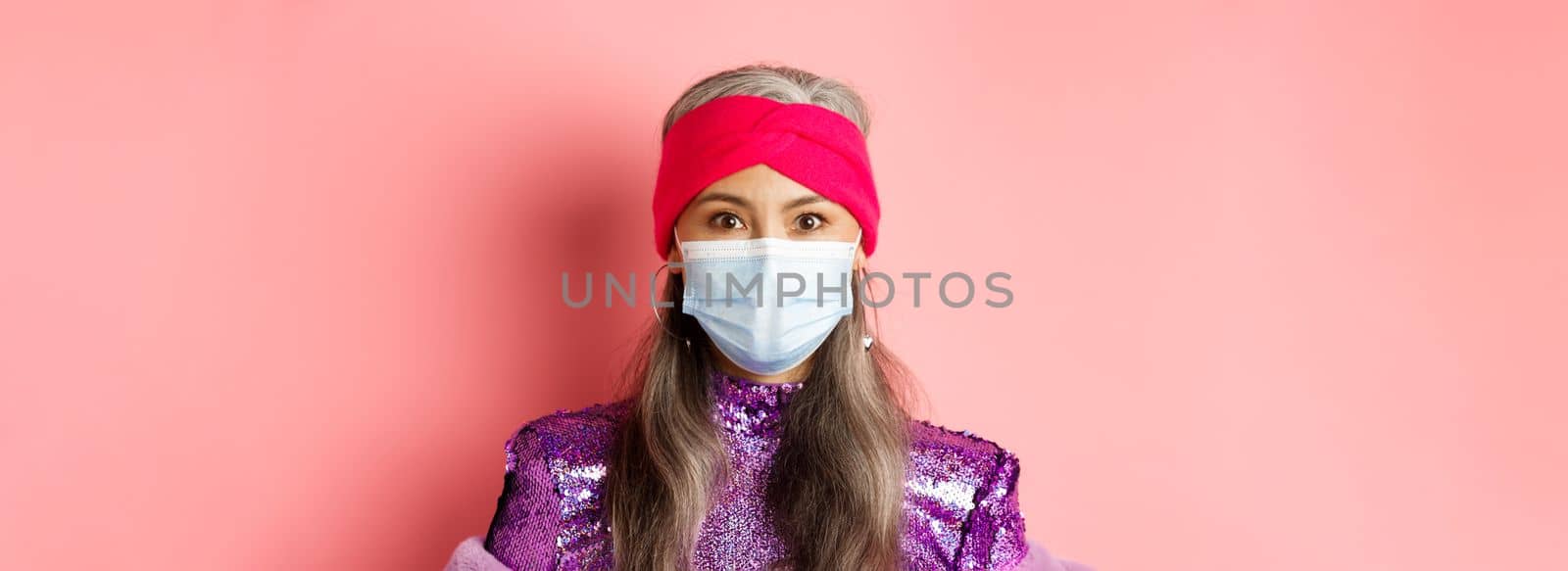 Covid-19, virus and social distancing concept. Close-up of stylish asian senior woman in medical mask and shiny dress looking at camera, standing over pink background by Benzoix