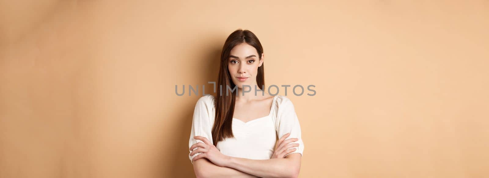 Young caucasian woman looking determined and confident, standing with arms crossed, smiling at camera, wearing white dress, beige background.