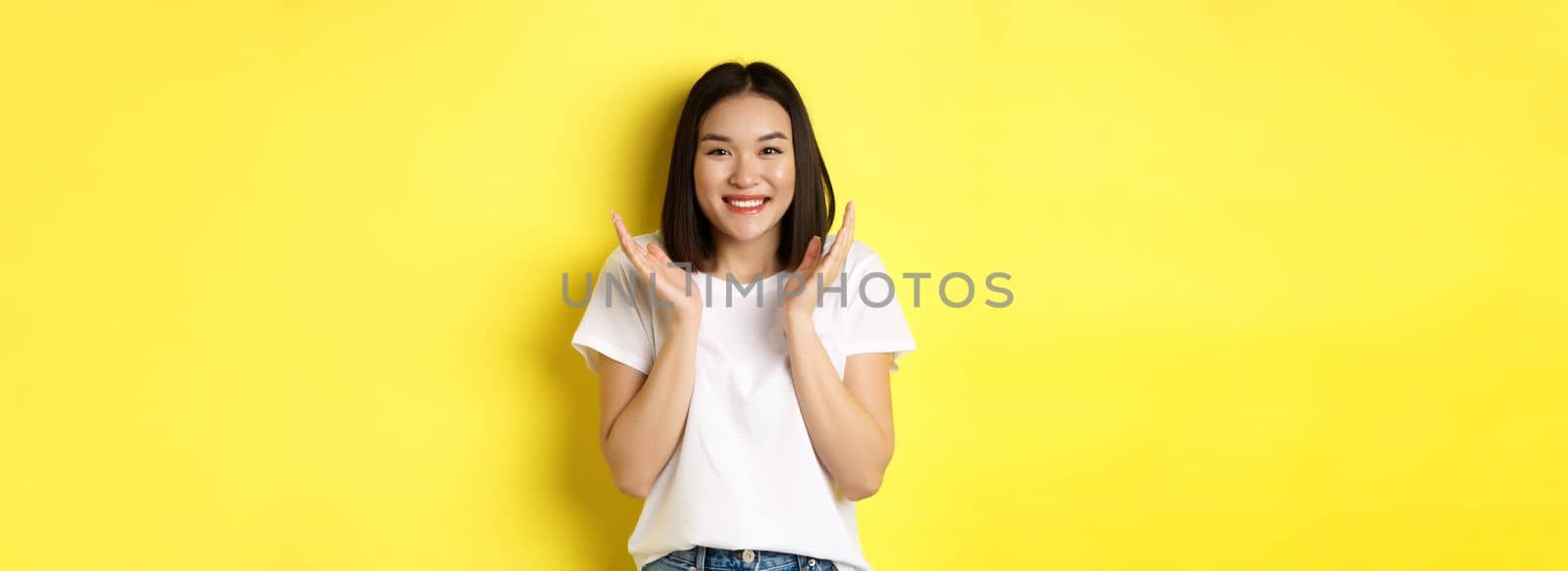 Beauty and fashion concept. Excited asian woman clap hands and smiling happy at camera, standing in white t-shirt against yellow background.