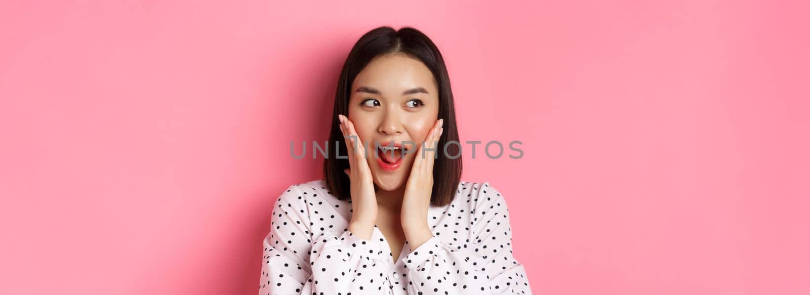 Close-up of beautiful asian woman looking surprised and excited, hear amazing news, looking left and rejoicing, standing against pink background.