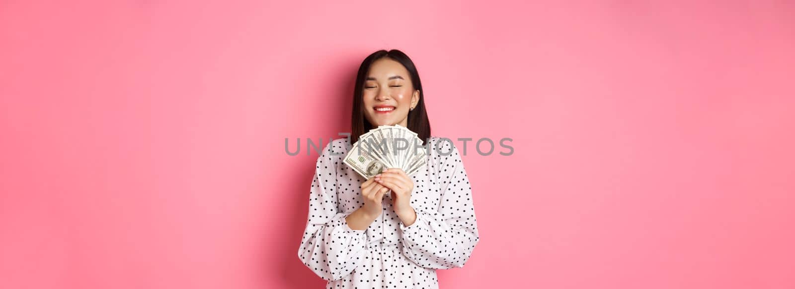 Shopping concept. Happy and satisfied asian woman winning prize money, showing dollars and rejoicing, standing over pink background by Benzoix