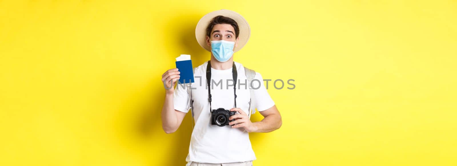 Concept of covid-19, travelling and quarantine. Happy man tourist with camera, showing passport and tickets for vacation, going on trip during pandemic, yellow background.