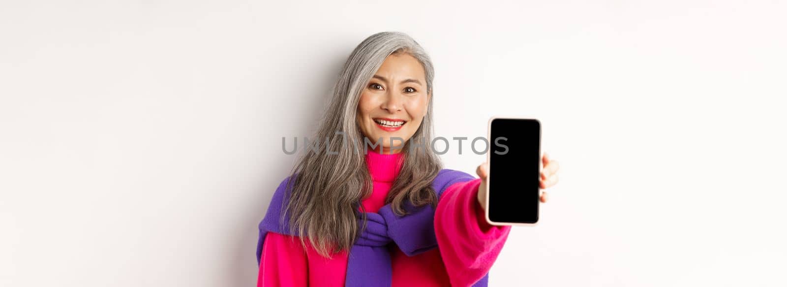 Online shopping. Close up of stylish asian senior woman extending hand with mobile phone, showing blank smartphone screen and smiling, standing over white background.