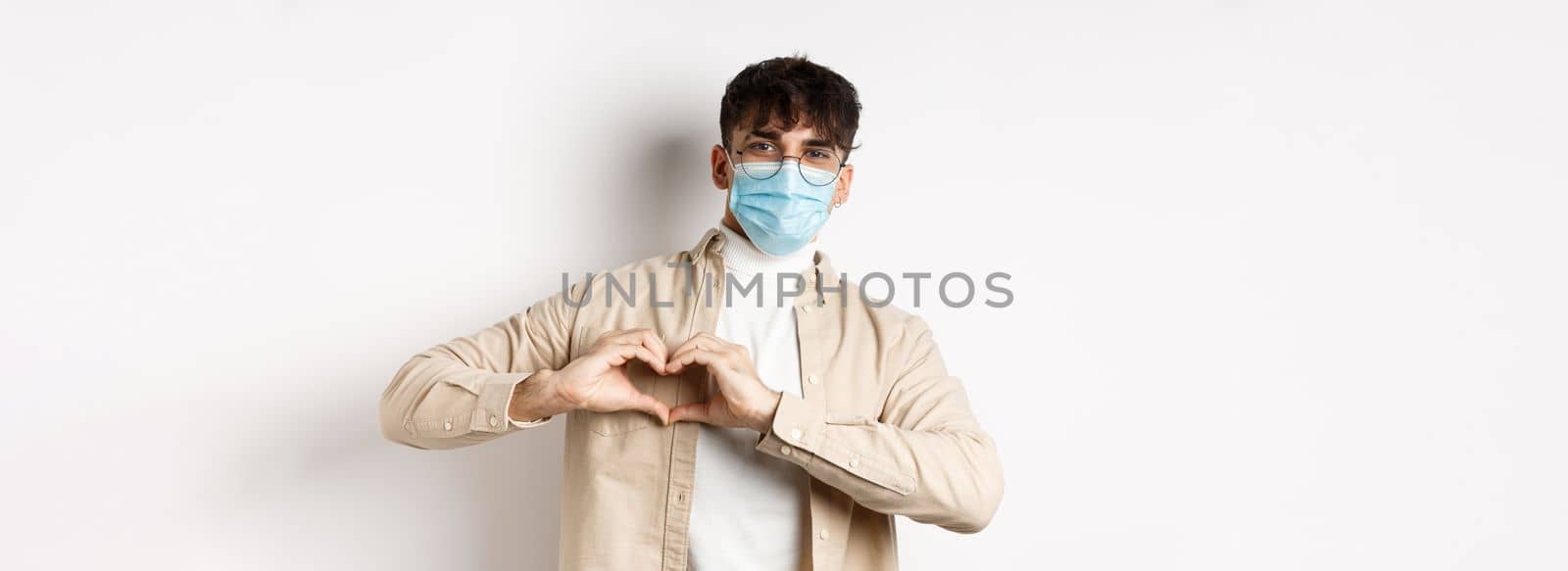 Health, covid and quarantine concept. Romantic young man in sterile medical mask showing heart gesture on chest, say I love you, standing on white background.