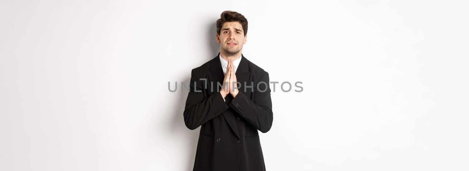 Image of handsome businessman in suit need help, begging you, holding hands in plead gesture and looking desperate, standing over white background.