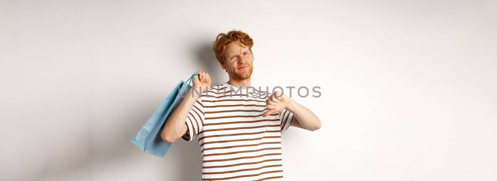 Disappointed young man with red hair and beard showing thumbs-down after bad shopping experience, holding bag over shoulder and frowning upset, white background.