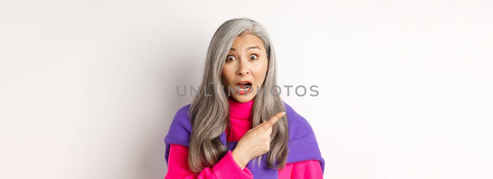 Close-up of amazed and intrigued asian senior woman pointing finger left at something cool, staring at camera impressed, standing over white background.