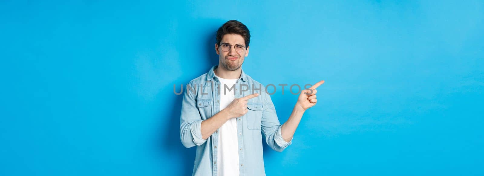 Displeased and skeptical male model in glasses, pointing fingers left at something bad, showing awful advertisement, standing over blue background by Benzoix