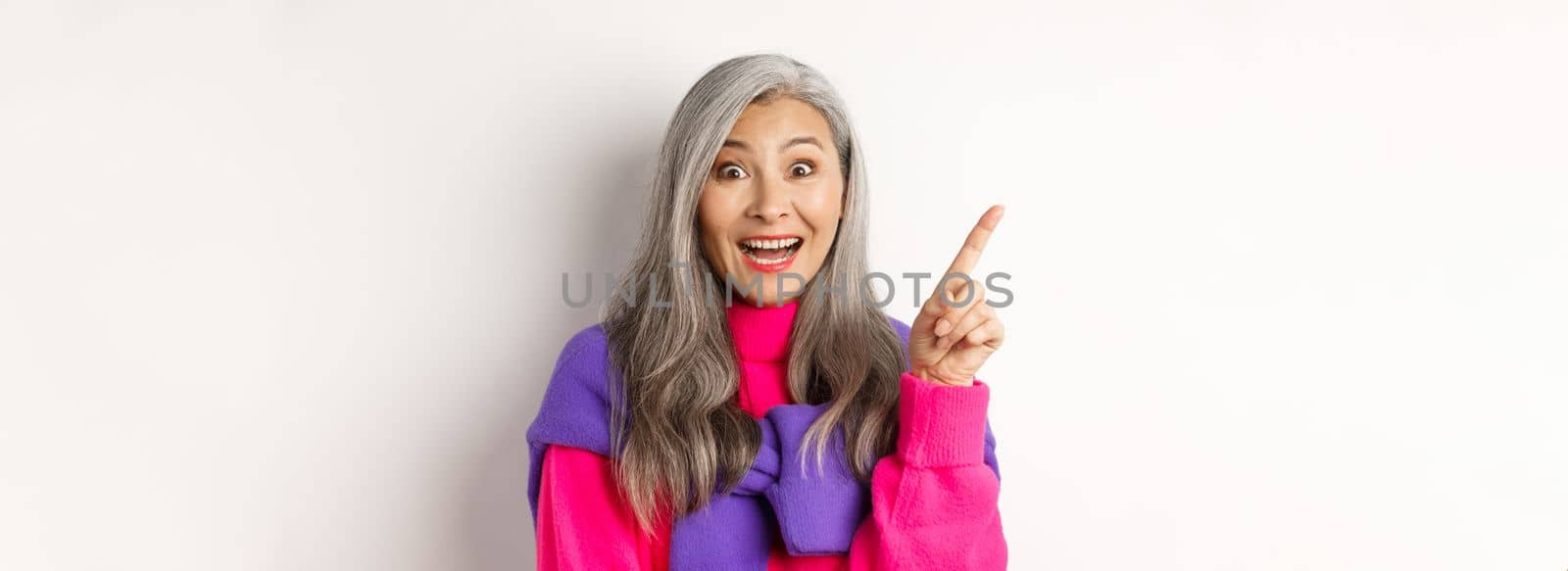 Close up of beautiful asian female model pointing finger at upper left corner, smiling and looking happy at camera, showing promo deal, white background.