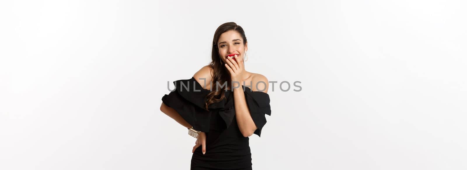 Fashion and beauty. Coquettish young woman in black dress, laughing and covering mouth with hand, posing flirty over white background.
