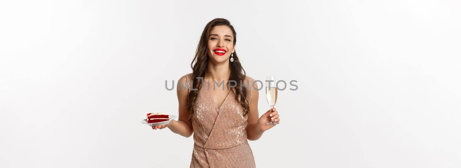 Party and celebration concept. Elegant woman with red lips, glamour dress, drinking champagne and eating cake, standing over white background.
