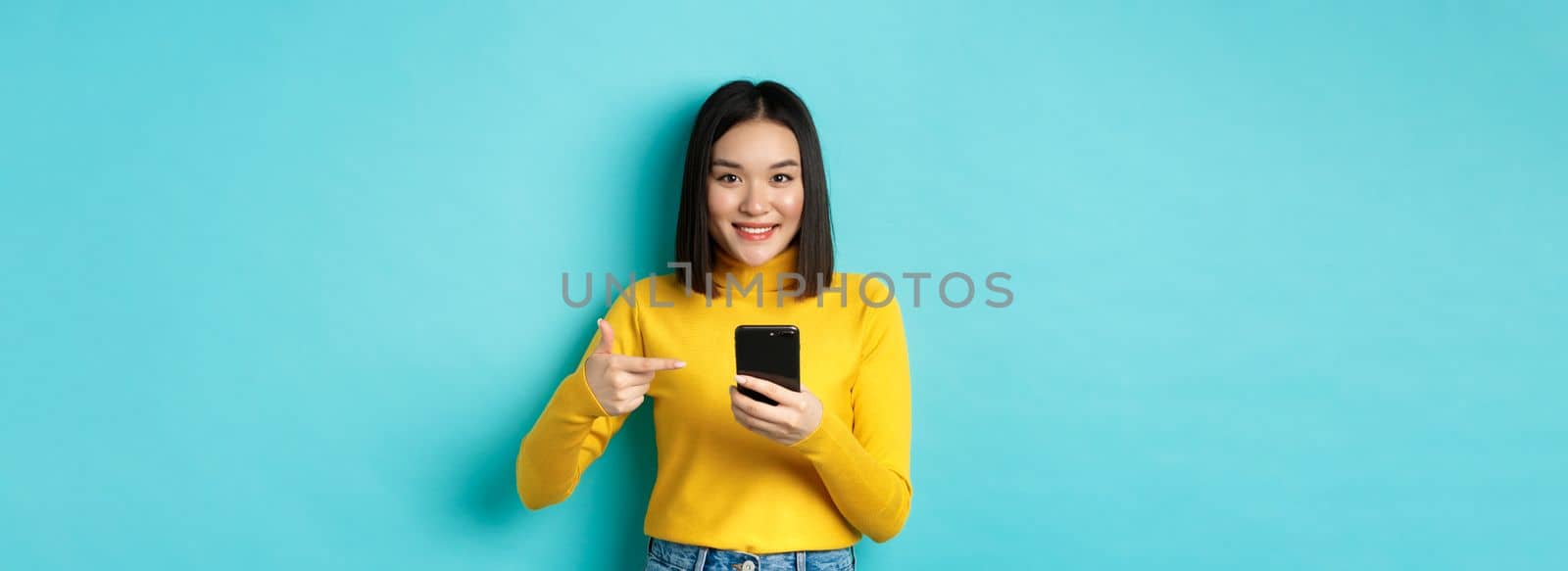 E-commerce and online shopping concept. Cute asian woman in yellow sweater pointing at smartphone, smiling at camera, standing over blue background.