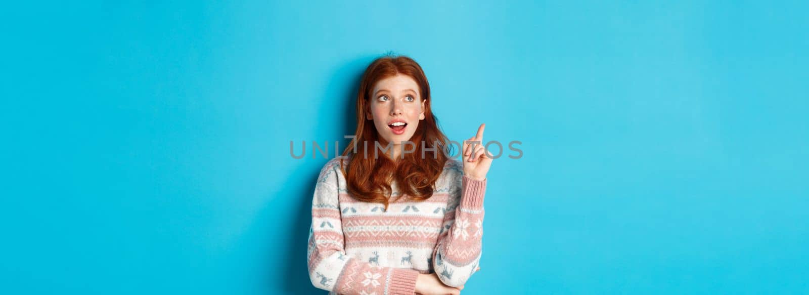 Thoughtful redhead girl in winter sweater having an idea, raising finger in eureka sign and looking up, standing against blue background.