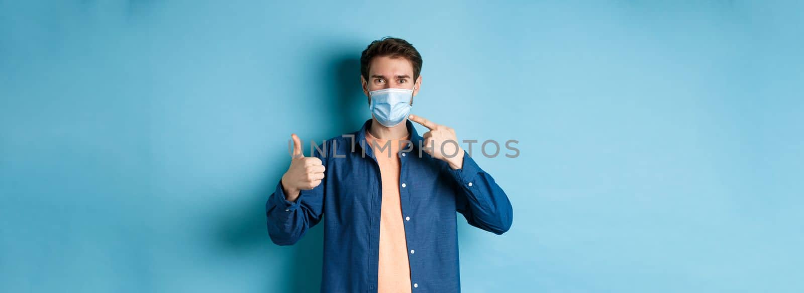 Coronavirus and pandemic concept. Young healthy man pointing at medical mask and showing thumbs up, using preventive measures from catching covid-19, blue background by Benzoix