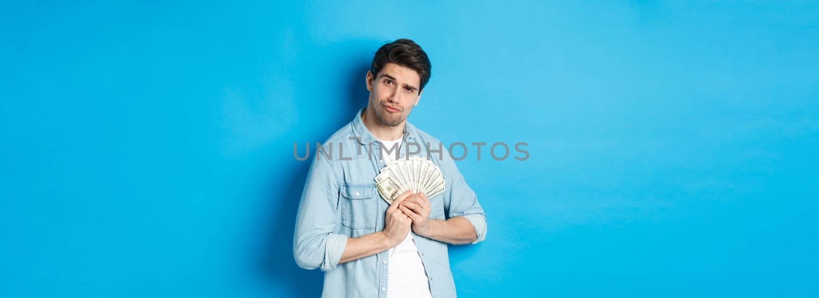 Handsome young man keeping money to himself, smiling and looking greedy, standing over blue background by Benzoix