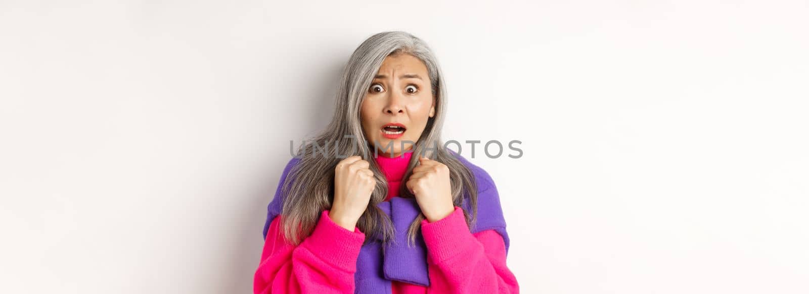 Close up of scared asian senior woman gasping, looking frightened and startled at camera, standing over white background by Benzoix