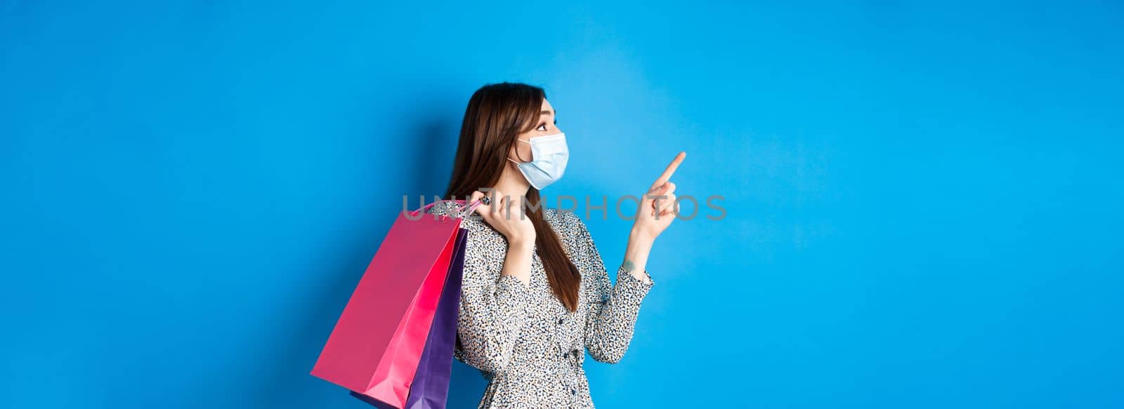 Covid-19, pandemic and lifestyle concept. Profile of excited girl wears medical mask on shopping, pointing and looking at upper left corner logo, holding purchases over shoulder, blue background.
