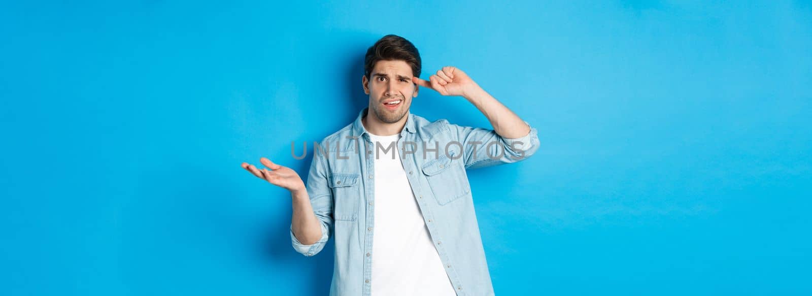 Confused man scolding person for being stupid, pointing finger at head and shrugging, looking puzzled, disapprove actions, standing against blue background.