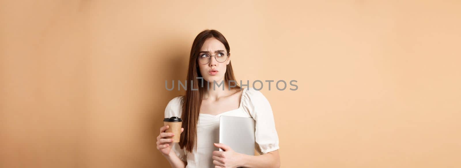 Confused and pensive girl with laptop looking aside suspicious, drinking takeaway coffee, standing against beige background.