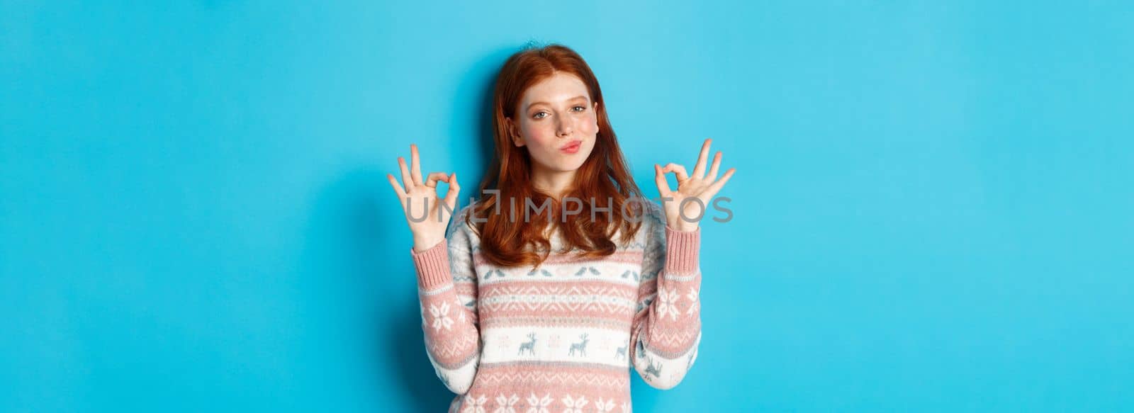 Satisfied and proud redhead girl nod in approval, showing okay sign, not bad or praise gesture, standing against blue background.