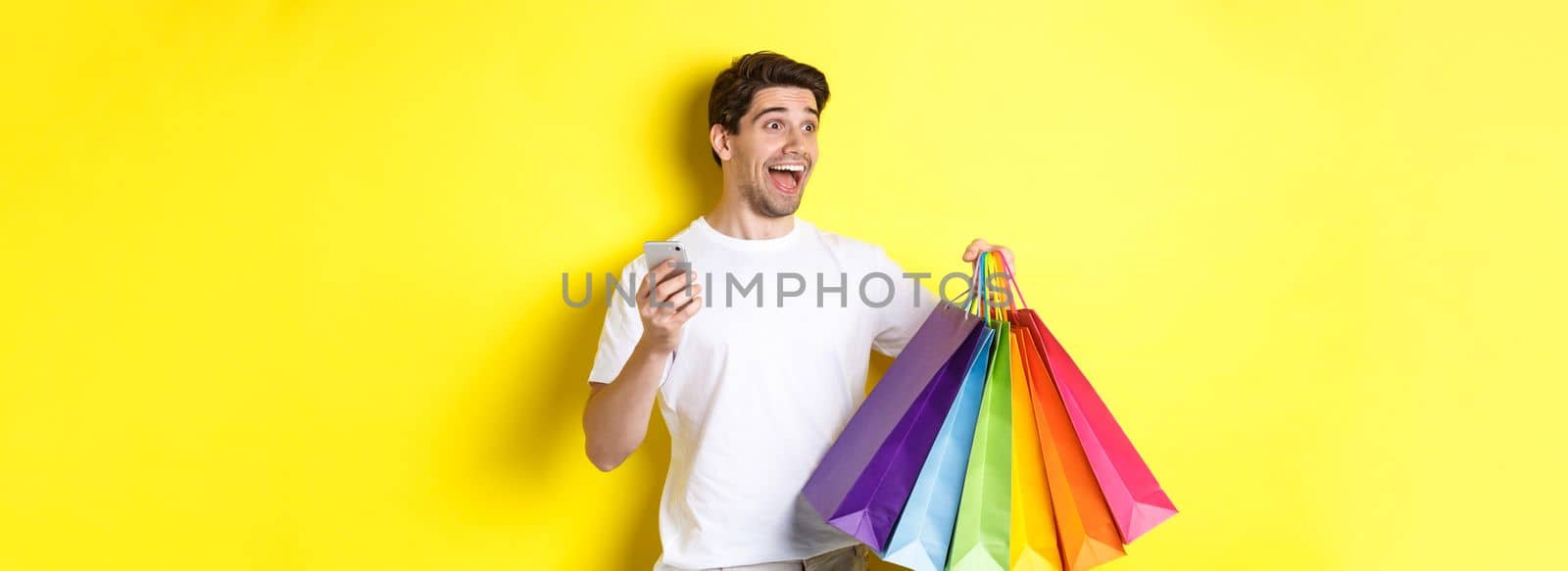 Concept of mobile banking and cashback. Happy man looking amazed, holding shopping bags and smartphone, yellow background.