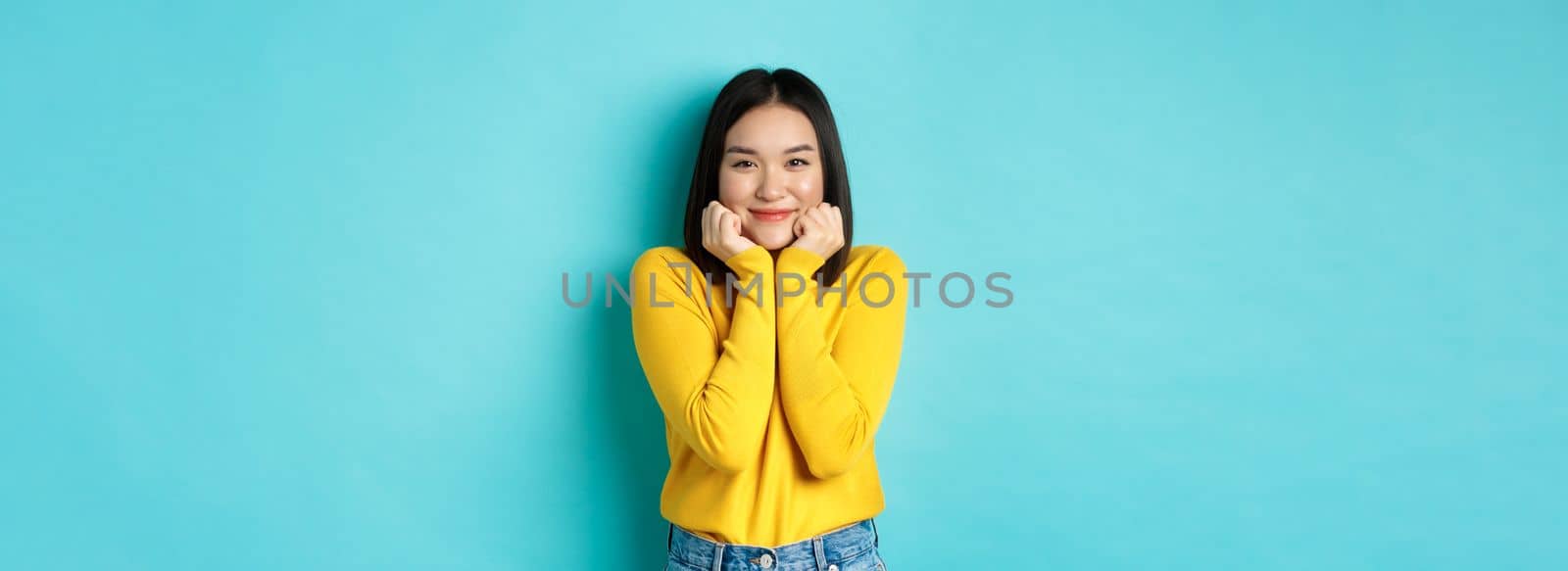 Beauty and fashion concept. Beautiful asian woman blushing and smiling, looking at something cute and silly, lean face on palms, standing over blue background.