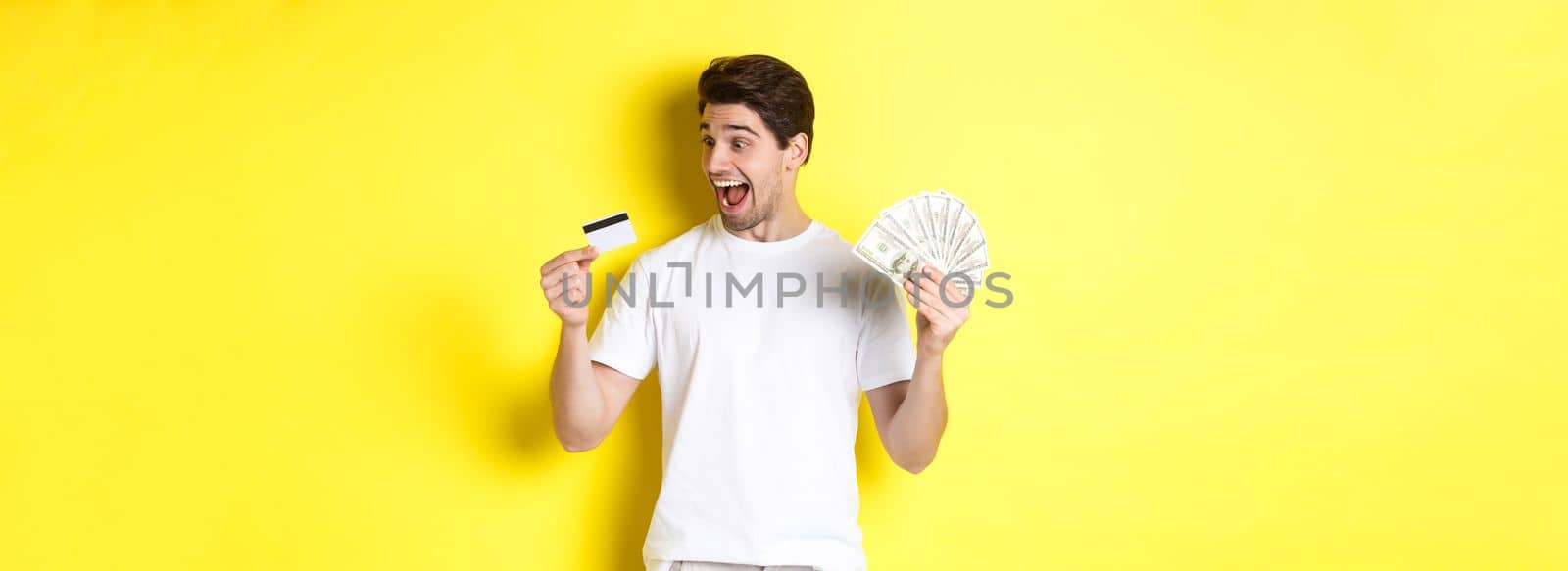 Cheerful guy looking at credit card, holding money, concept of bank credit and loans, standing over yellow background.