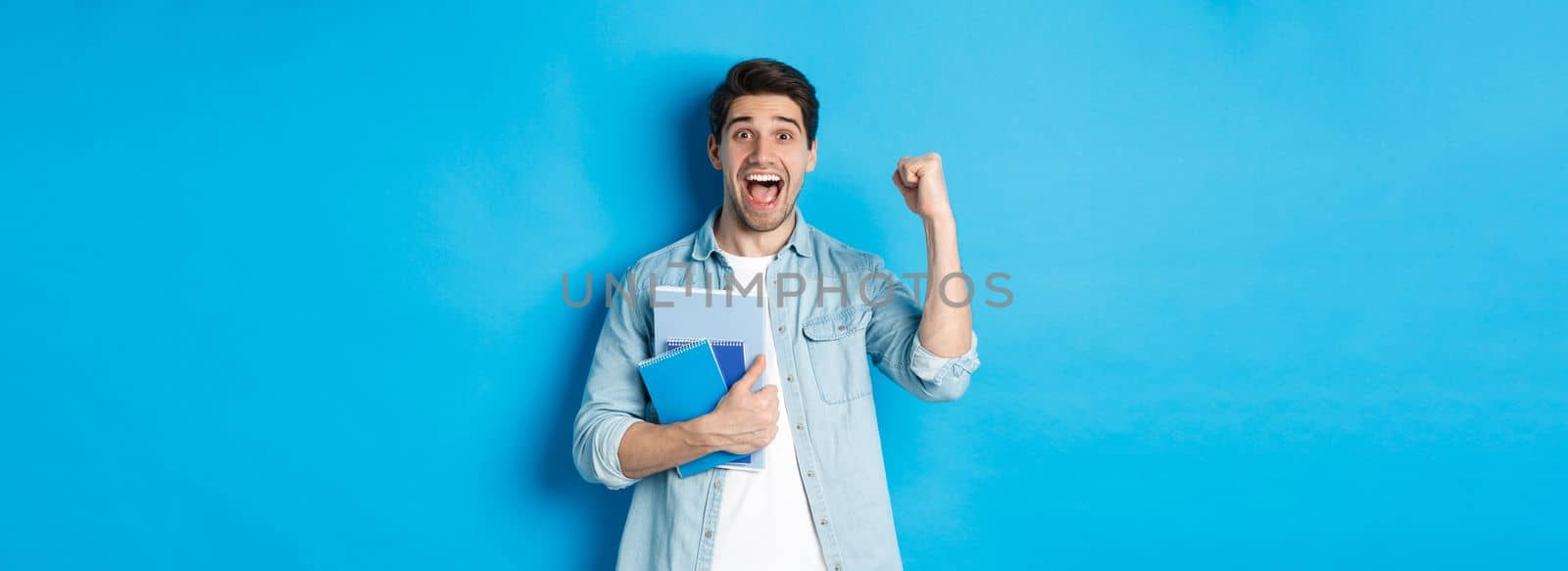 Cheerful guy holding notebooks and celebrating, making fist pump and shouting yes with excitement, standing over blue background.