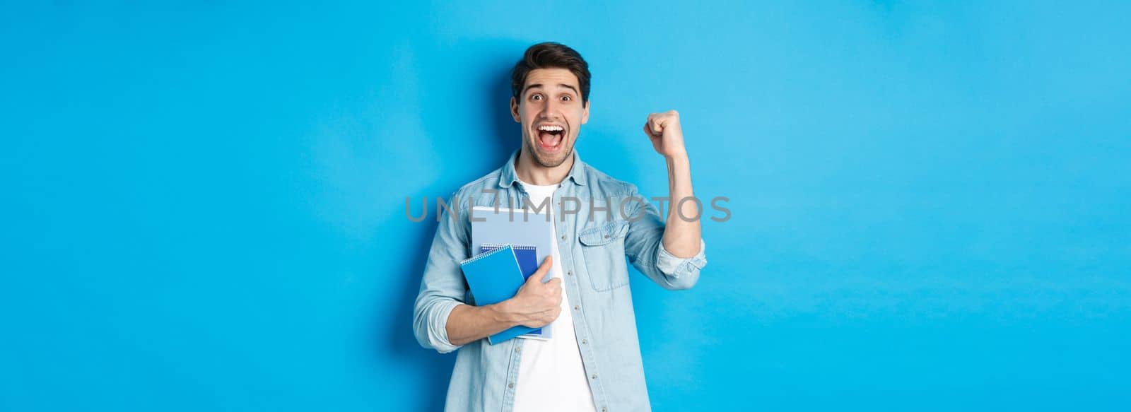 Cheerful guy holding notebooks and celebrating, making fist pump and shouting yes with excitement, standing over blue background by Benzoix