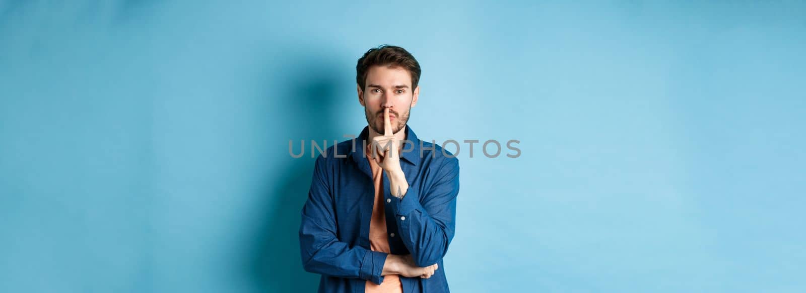 Modern guy shushing at camera, press finger to lips and telling a secret, making taboo sign, standing on blue background.