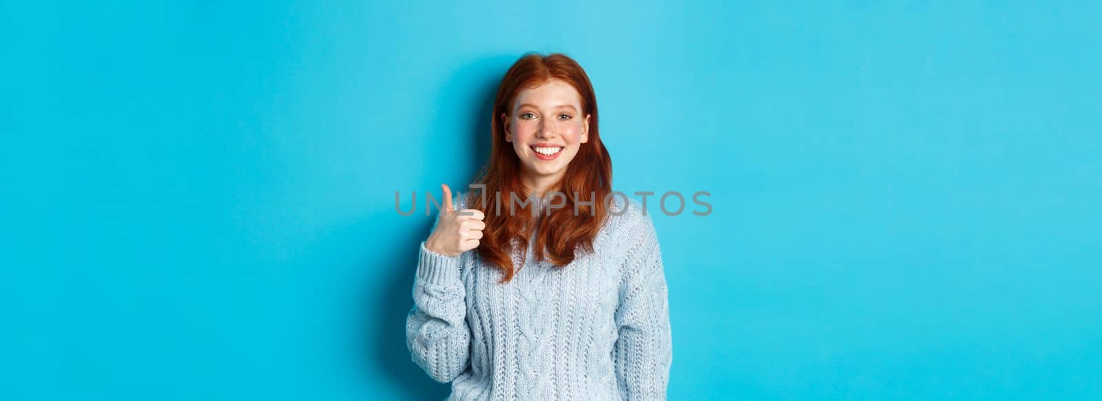 Cute redhead girl in sweater showing thumb up, like and agree, smiling pleased, standing against blue background.