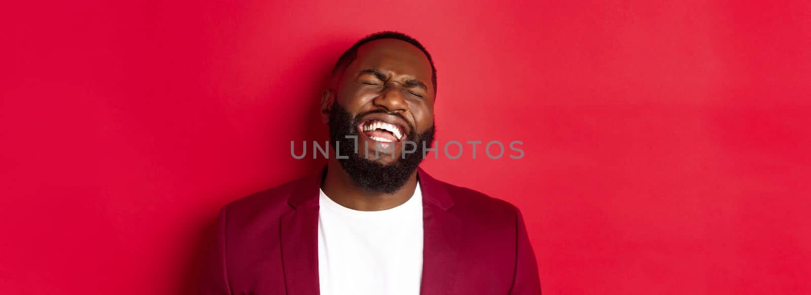 Close-up of happy and carefree Black man having fun, laughing and smiling, standing in blazer against red background.