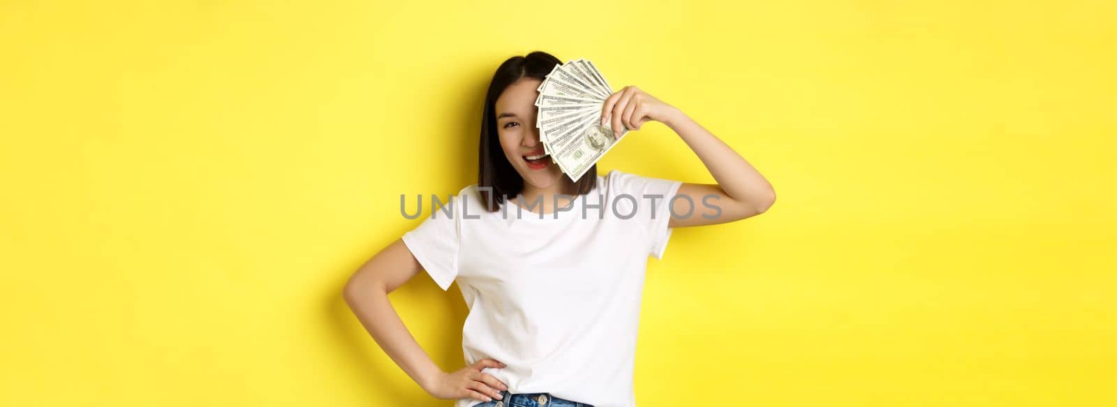 Cute asian woman hiding face behind money, peeking at camera satisfied, earn cash, standing over yellow background.