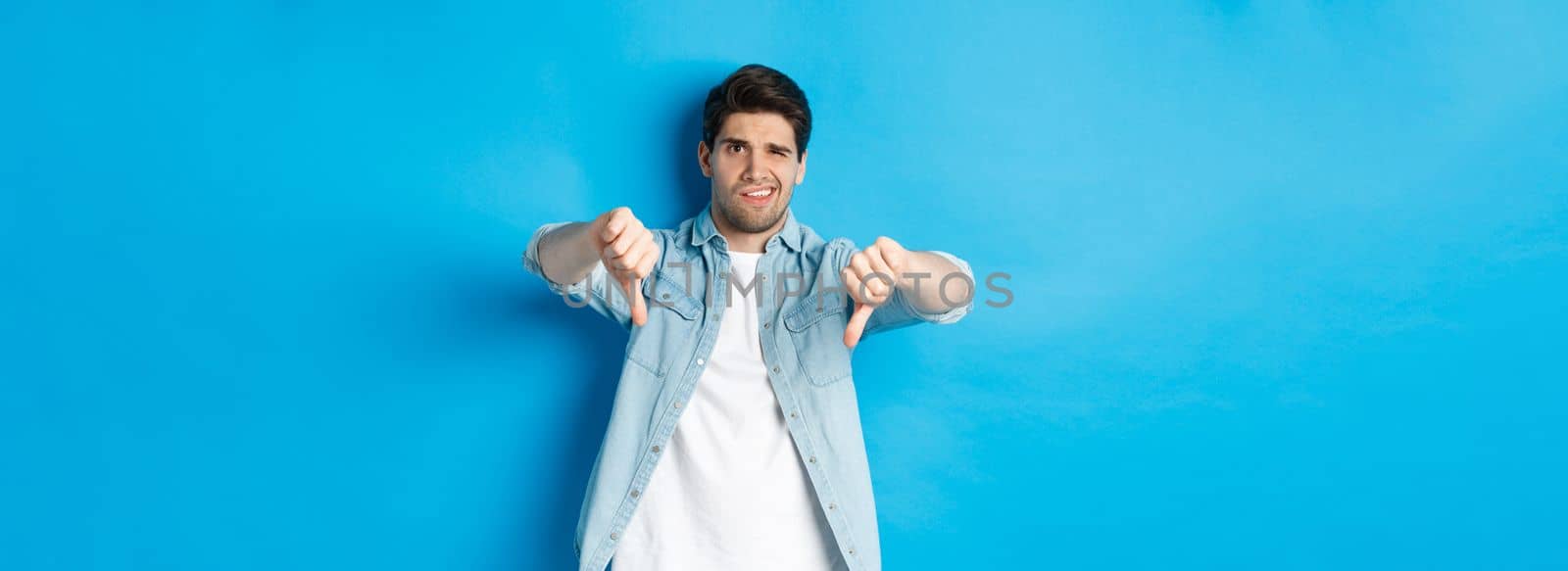 Disappointed man showing thumbs down and grimacing at something awful, dislike product, judging bad advertisement, standing against blue background.