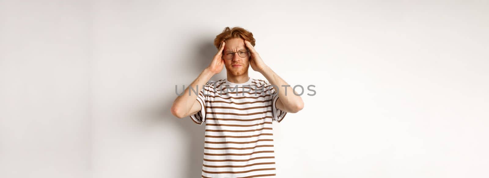 Image of young man with red hair and glasses touching head, frowning from painful migraine, having headache, standing over white background.