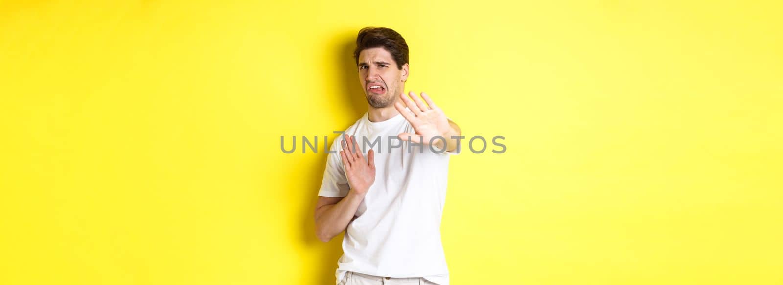 Disgusted man refusing, grimacing from dislike and aversion, begging to stop, standing in white t-shirt against yellow background.