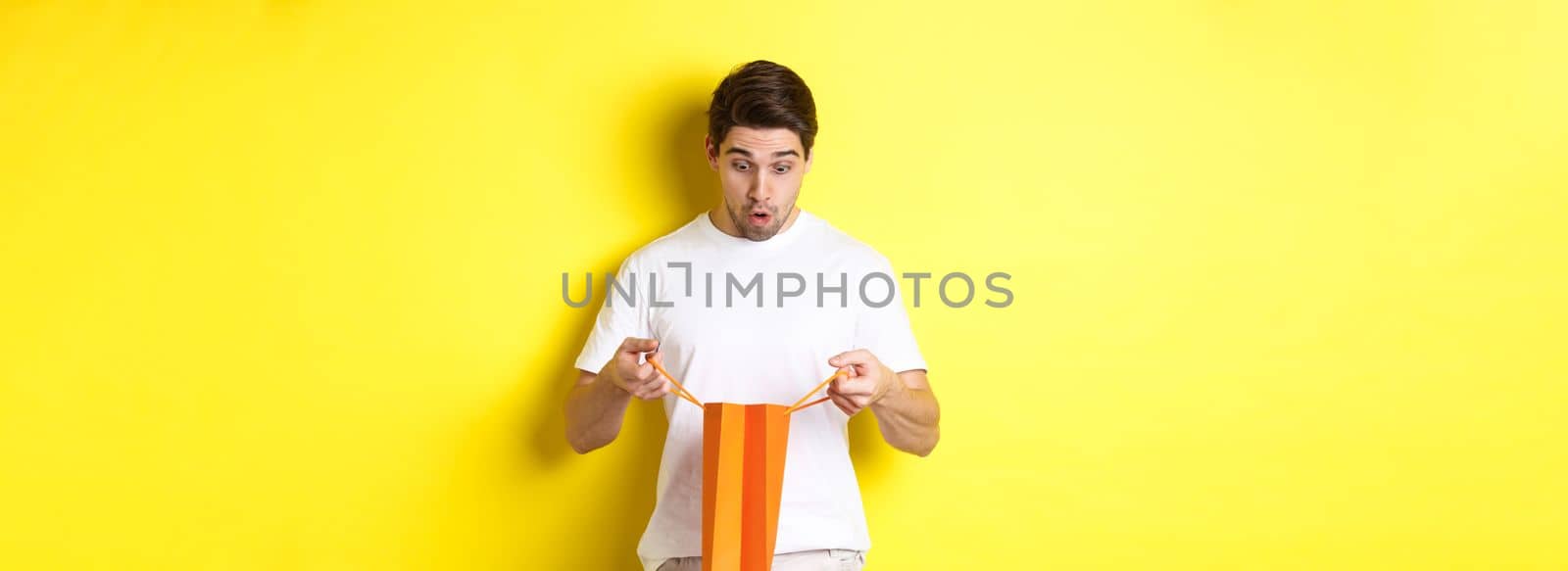 Surprised boyfriend open bag with gift, looking inside amazed, standing over yellow background by Benzoix