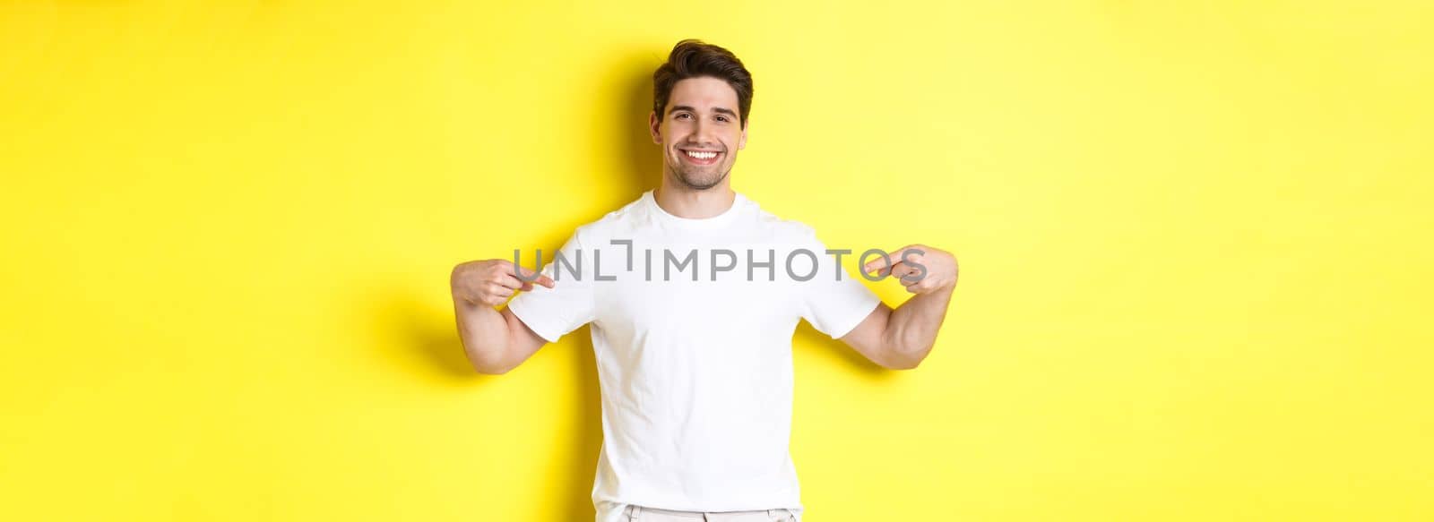 Happy attractive guy pointing fingers at your logo, showing promo on his t-shirt, standing over yellow background.