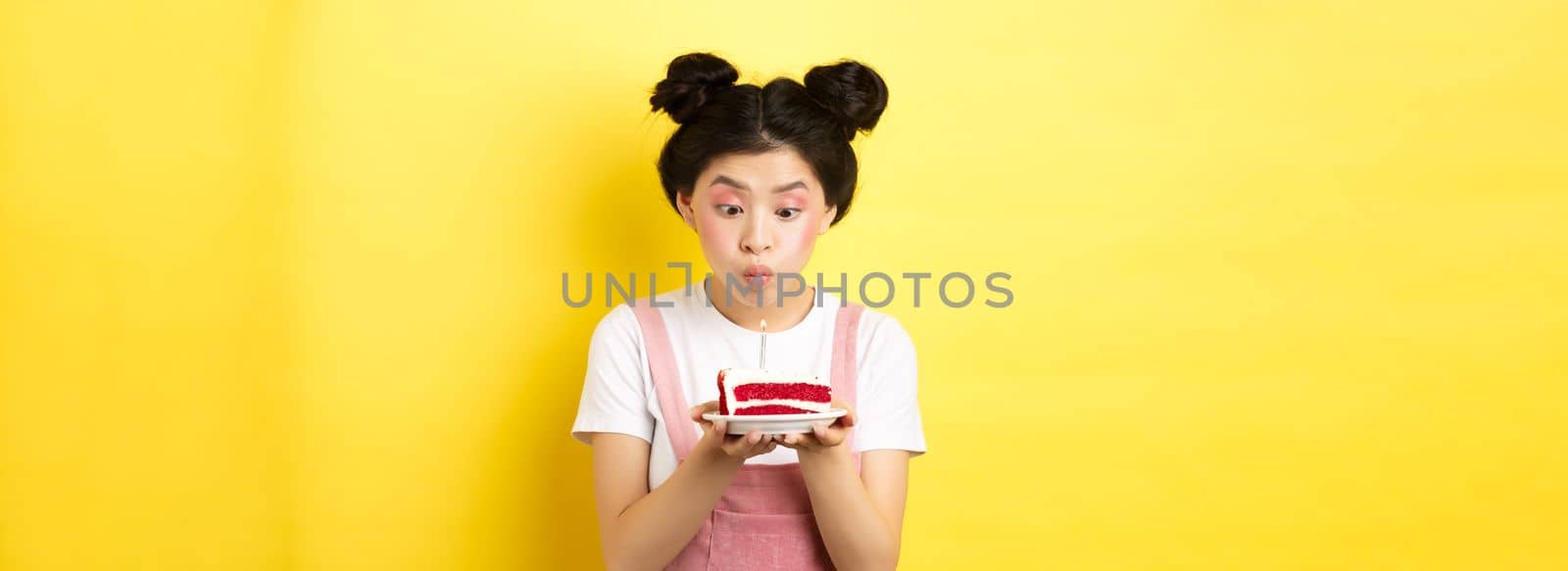 Holidays and celebration. Silly asian girl with glamour makeup, making wish and blowing candle on birthday cake, standing on yellow background.