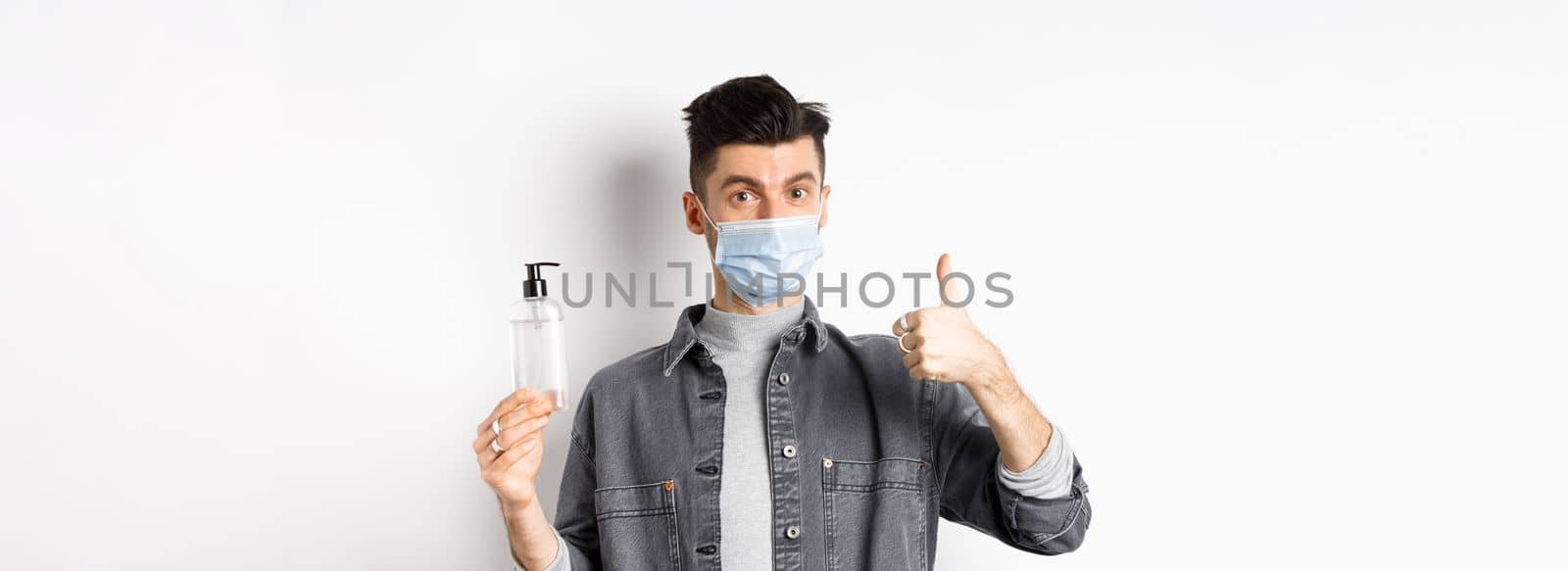 Healthy people and covid-19 concept. Excited man in sterile medical mask holding bottle of good hand sanitizer, show thumb up, recommend antiseptic, standing against white background by Benzoix