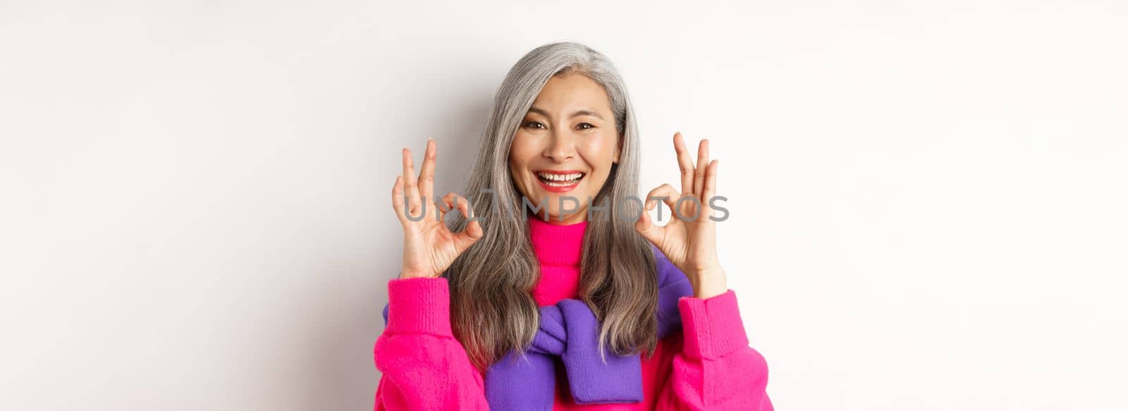 Close-up of satisfied asian female with grey hair, smiling pleased and showing OK signs, approve and like product, praising something awesome, white background by Benzoix