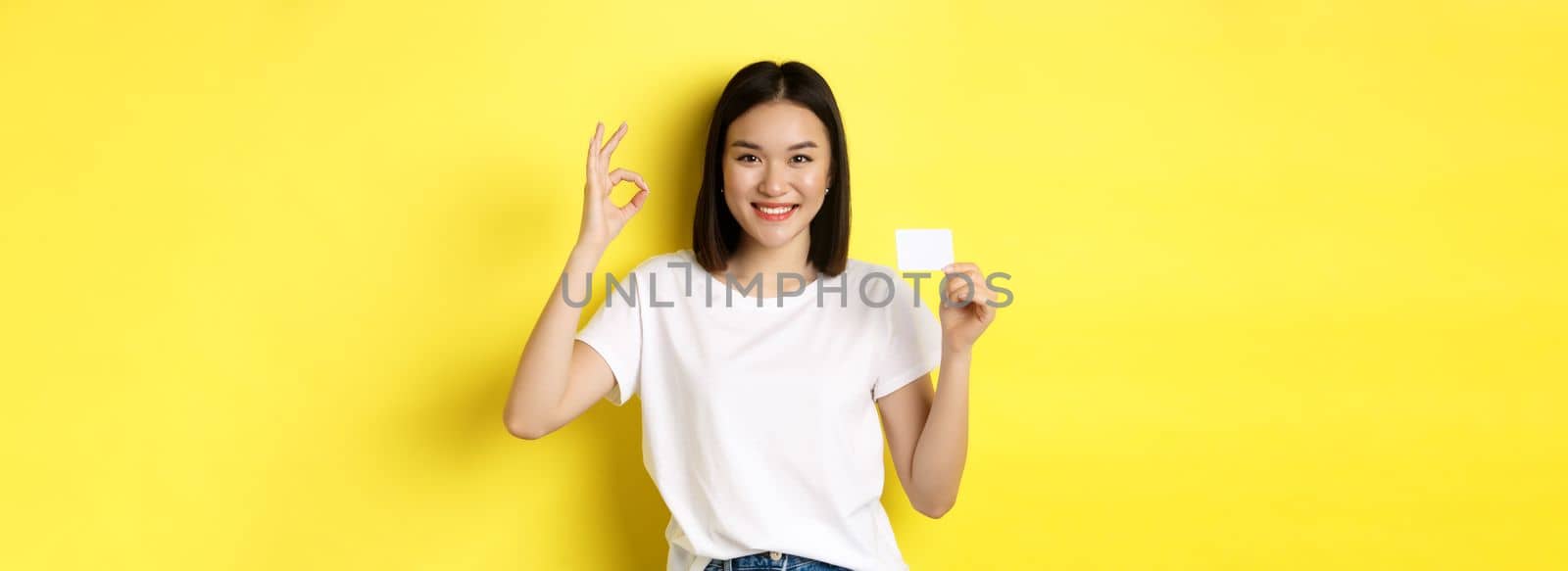 Young asian woman in casual white t-shirt showing plastic credit card and okay gesture, recommend bank, smiling at camera, yellow background.