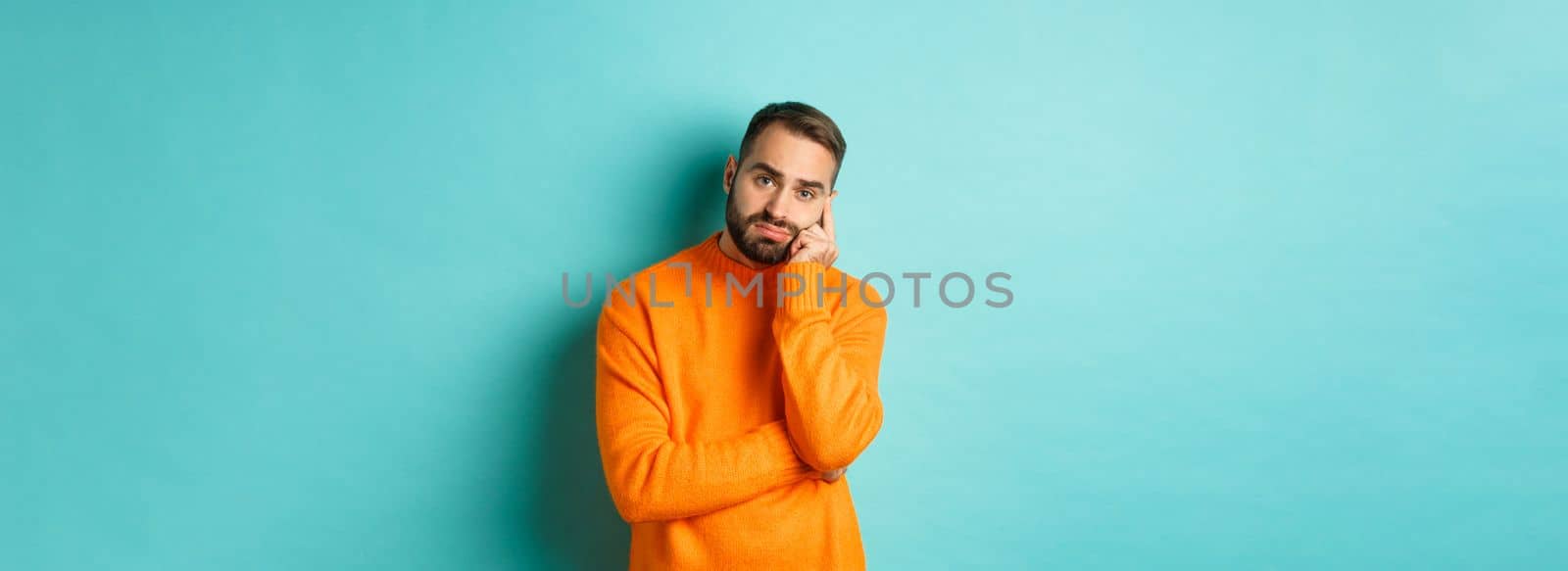 Sad and gloomy man with beard, looking indesicive and sad, thinking about something and sulking, standing over turquoise background.