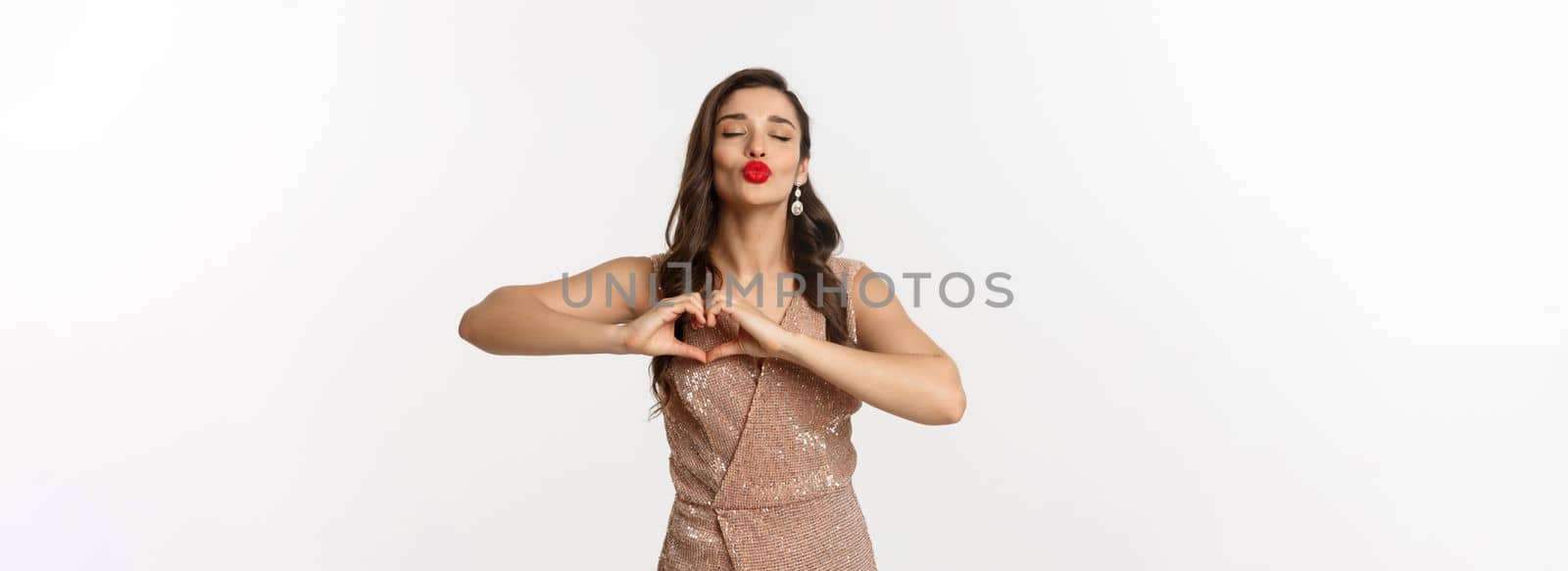 Celebration and party concept. Beautiful young woman in stylish dress, red lips, showing heart sign and waiting for kiss, standing over white background.