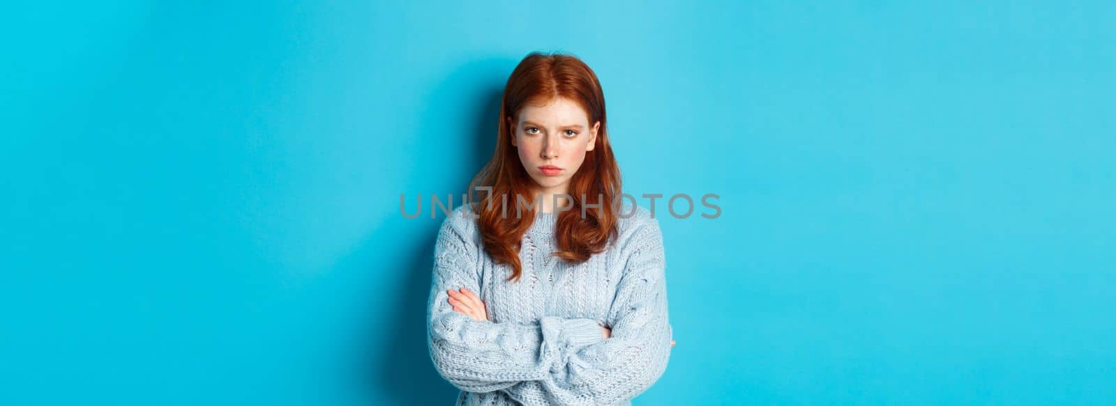 Image of angry redhead girl feeling offended, cross arms on chest and sulking, staring at camera mad, standing against blue background by Benzoix