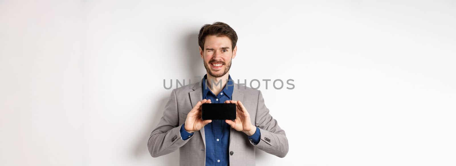 E-commerce and online shopping concept. Cheeky male model in suit winking and showing empty smartphone screen, smiling happy at camera, white background.