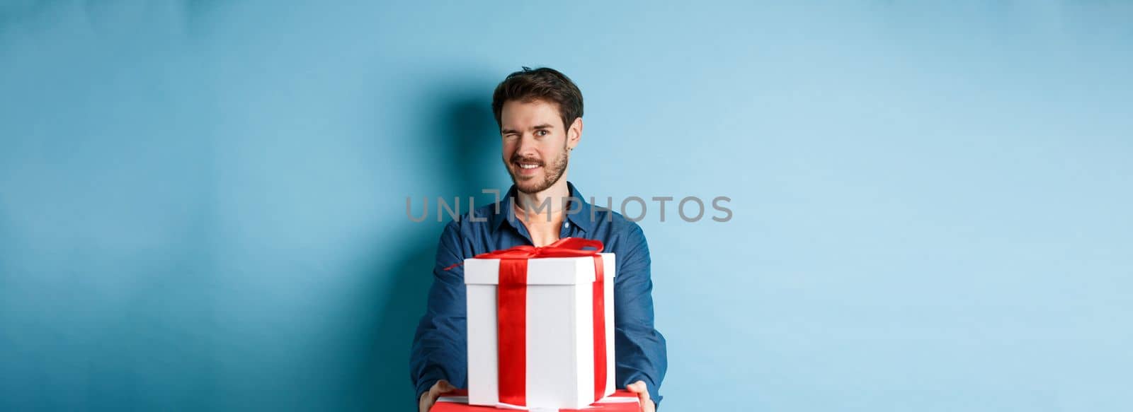 Handsome boyfriend winking at camera and wishing happy valentines day, extending hands with gift boxes, making romantic surprise, standing over blue background by Benzoix