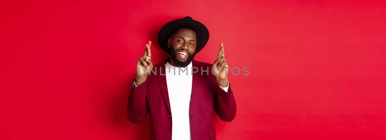 Hopeful african american man making wish, holding fingers crossed for good luck and smiling optimistic, standing against red party background.